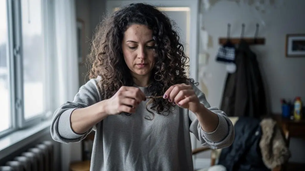 Une femme aux cheveux bouclés étire une mèche entre ses doigts près d'une fenêtre lumineuse pour observer la forme de sa boucle.